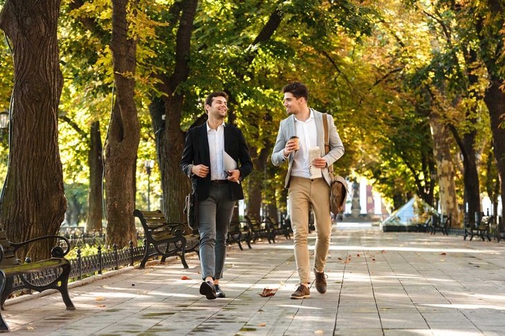 Photo of young businessmen in suits walking outdoor through green park with takeaway coffee and laptop during sunny day